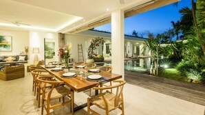 A modern dining area with a wooden table and chairs, a sofa, and a view of a pool and garden at dusk. - Villa Hugo (Canggu)