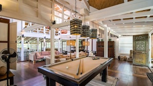 Une table de billard se trouve dans une pièce spacieuse, dotée de hauts plafonds et de poutres en bois, et est entourée de sièges confortables et de lanternes suspendues.