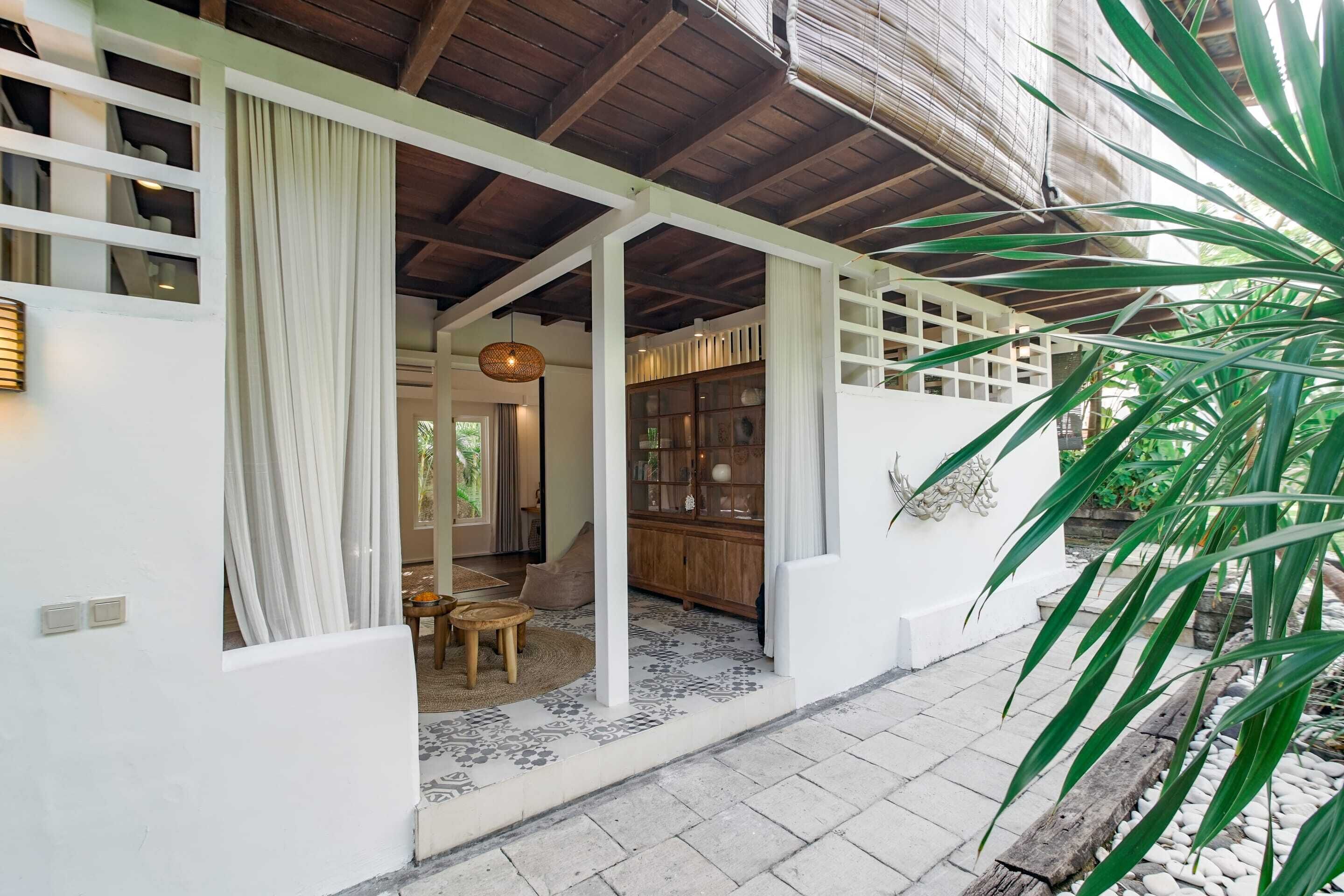 A covered outdoor area with a wooden ceiling, white walls, and a patterned floor. There is a wooden table and chairs, a hanging light fixture, and a glass cabinet with decorative items.
