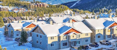 Una zona residencial nevada con casas modernas, un estacionamiento y montañas al fondo.