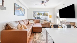 A modern living room with a sectional sofa, a dining table, and a large TV. - Cozy Condo Located  Minutes from the Biggest Skiing in America (Big Sky)