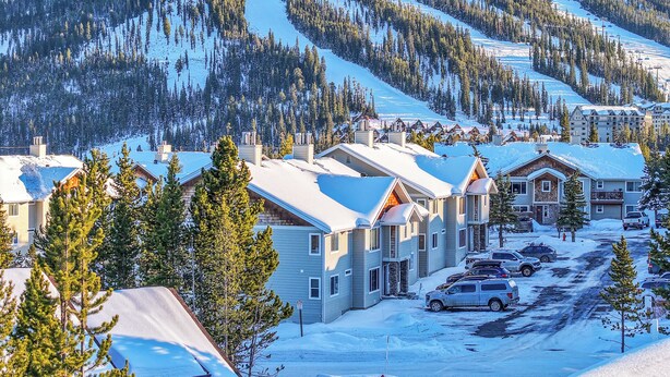 A snow-covered mountain resort with multiple buildings, including a large lodge and smaller cabins, surrounded by dense forests and ski slopes. - Cozy Condo Located  Minutes from the Biggest Skiing in America (Big Sky)