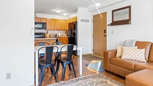 A modern kitchen with wooden cabinets, a white countertop, and a dining area with a white table and black chairs. There is a brown leather sofa with patterned pillows in the living area. - Cozy Condo Located  Minutes from the Biggest Skiing in America (Big Sky)