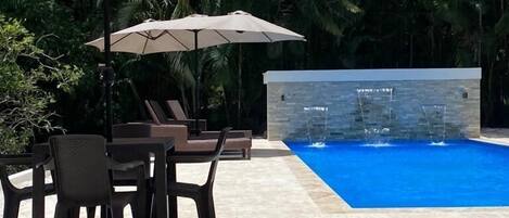 A pool area with stone flooring, two umbrellas, and lounge chairs.