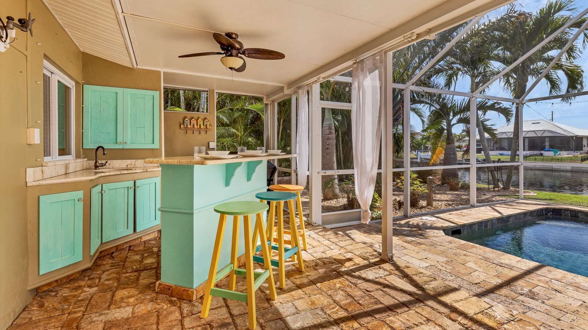 Een keuken met turquoise kastjes, een bar met krukken, een plafondventilator en uitzicht op een zwembad en palmbomen.