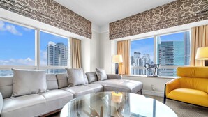 Een moderne hotelkamer met een grijze bank, een gele fauteuil, een glazen bijzettafeltje en grote ramen met uitzicht op de stad.