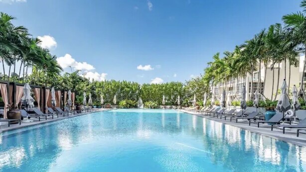 Ein großer, klarer Pool mit Liegestühlen und Sonnenschirmen an beiden Seiten, umgeben von Palmen und blauem Himmel.
