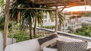 Ein weißes Outdoor-Sofa mit gemusterten Kissen unter einer Holz-Pergola, umgeben von Grün und mit Blick auf eine Stadtlandschaft.