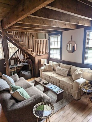 A cozy living room with a wooden staircase, a beige sofa, a coffee table, and a small side table. - Cozy Quechee Cabin near Woodstock and Skiing w/woodstove and all the amenities (Hartford)