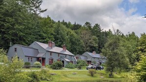 Une rangée de maisons à deux étages, avec un étang au premier plan, le tout entouré d’une forêt dense.