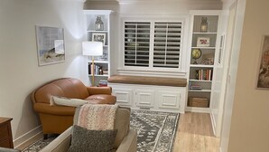 A living room with a brown leather sofa, a beige armchair, and a patterned rug. There is a white built-in bench with a window above it, and a bookshelf filled with books and decorative items. - Beachy Home in Provo. Large deck with builtin hot tub, Fire pit, Palapa (Provo)