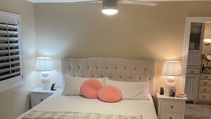 A neatly arranged bedroom with a tufted headboard, a bed with a white comforter and pink pillows, two bedside tables with lamps, a ceiling fan, and a mirror cabinet. - Beachy Home in Provo. Large deck with builtin hot tub, Fire pit, Palapa (Provo)