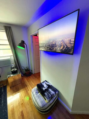 A room with a large flat-screen TV mounted on the wall, a standing floor lamp, a pet bed, and a small table with a lamp. - MANHATTAN BEST LOCATION! 2-BED Union Square and Village (New York)
