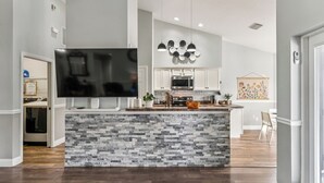 A modern kitchen with a stone backsplash, white cabinets, a large flat-screen TV, and pendant lighting. - TROPICAL RETREAT WITH PRIVATE HEATED POOL-TAMPA BAY. (New Port Richey)