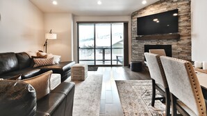 A modern living room with a leather sofa, a fireplace, a flat-screen TV mounted on a brick wall, and a large sliding glass door leading to a balcony.