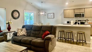 A modern living room with a gray sofa, a dining area with white chairs, a kitchen with wooden cabinets and a microwave, and a ceiling fan. - Cheerful  NEW Executive Home with office-Meridian (Meridian)