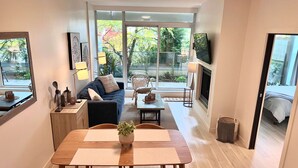 Ein modernes Wohnzimmer mit einer Couch, einem Esstisch, Stühlen, einem Kamin und großen Fenstern mit Blick auf Bäume.