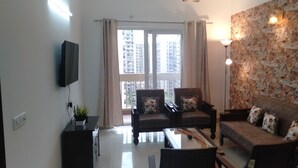 Ein Wohnzimmer mit einer Couch, Sesseln, einem Couchtisch, einem Fernseher und einem Balkon mit Blick auf Hochhäuser.