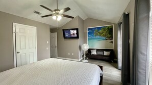 Una camera da letto con un letto, una ventola a soffitto, una televisione, un quadro raffigurante una spiaggia e una porta.