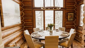 A cozy log cabin dining area with a round table set for six, surrounded by plush chairs, large windows with a snowy view, and a hanging light fixture. - Luxury Townhome-Bike/Hike-Out the Door Access-Private Hot Tub-3 King Bed En Suite-Bunk Room (Mountain Village)