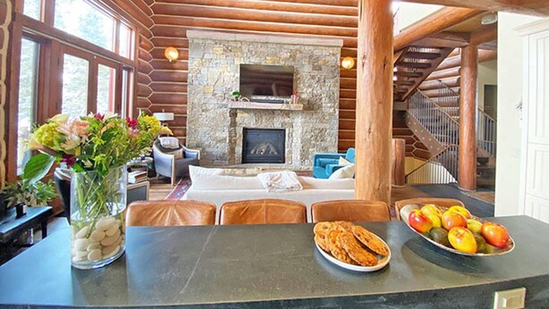 A cozy cabin interior with a stone fireplace, wooden beams, a dining area with a table and chairs, and a fruit bowl on the counter. - Luxury Townhome-Bike/Hike-Out the Door Access-Private Hot Tub-3 King Bed En Suite-Bunk Room (Mountain Village)