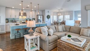 A spacious living area with a large sofa, a coffee table, and a side table with a lamp. There is a kitchen area with modern appliances and pendant lighting. - Brand New Pool! Incredible Ocean Views! Updated Beach House, Fenced Yard, Balcony, Easy Beach Access (St. Augustine)