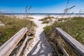 Un sentiero in legno conduce a una spiaggia con sabbia ed erba ai lati.
