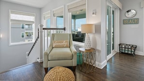 A spacious living room with a large window, a green armchair, a round ottoman, and a small side table with a lamp. - Brand New Pool! Incredible Ocean Views! Updated Beach House, Fenced Yard, Balcony, Easy Beach Access (St. Augustine)