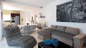 Ein modernes Wohnzimmer mit einer Couch, einem gemusterten Sessel, einem Essbereich mit Tisch und Stühlen sowie Blick auf die Stadt.