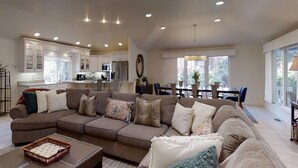 A spacious living room with a large sectional sofa, a dining area with a table and chairs, a kitchen with white cabinets, and a large window providing natural light.