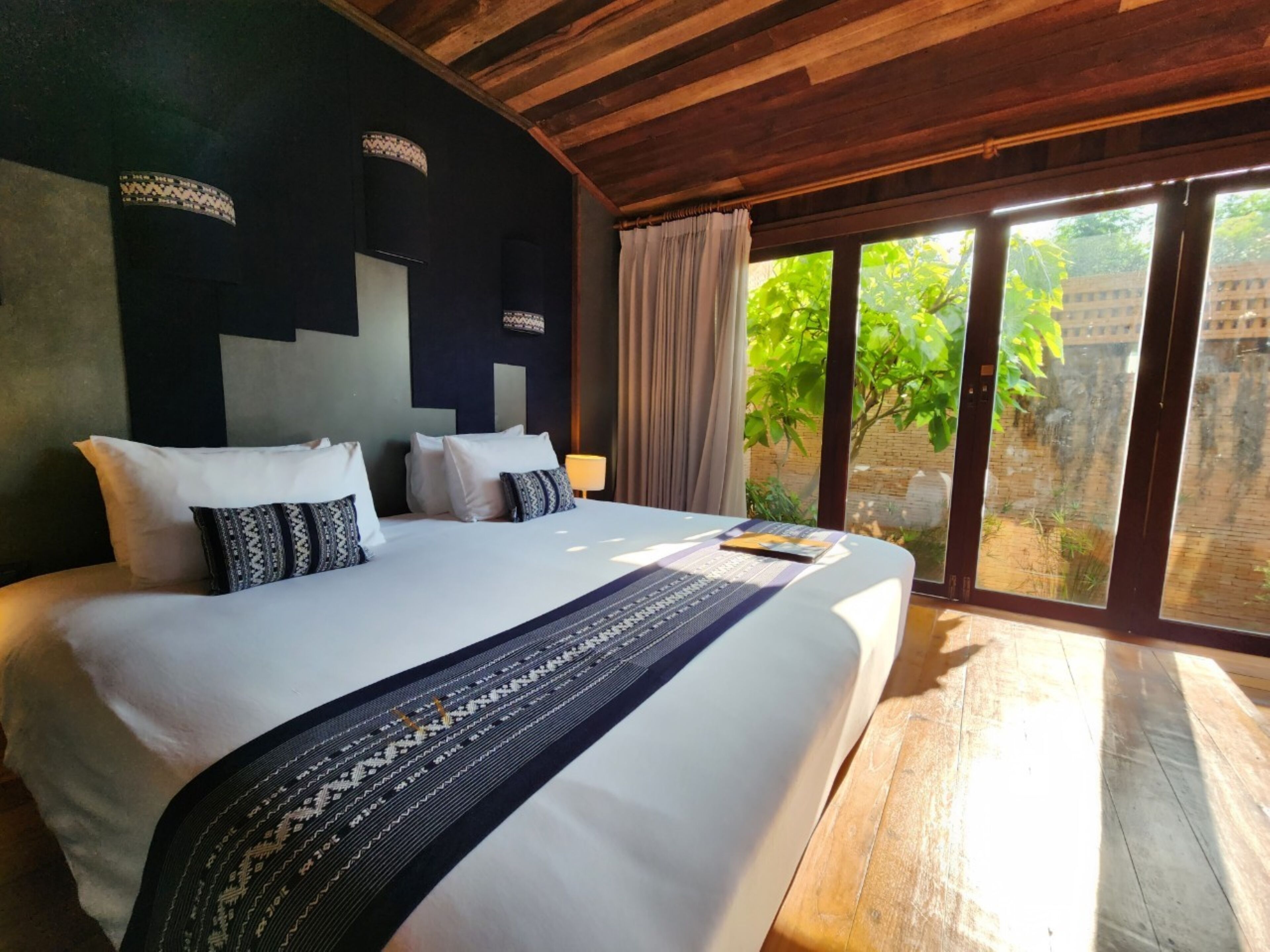 A large bed with white linens and patterned pillows, a wooden ceiling, and a view of greenery through a sliding door.
