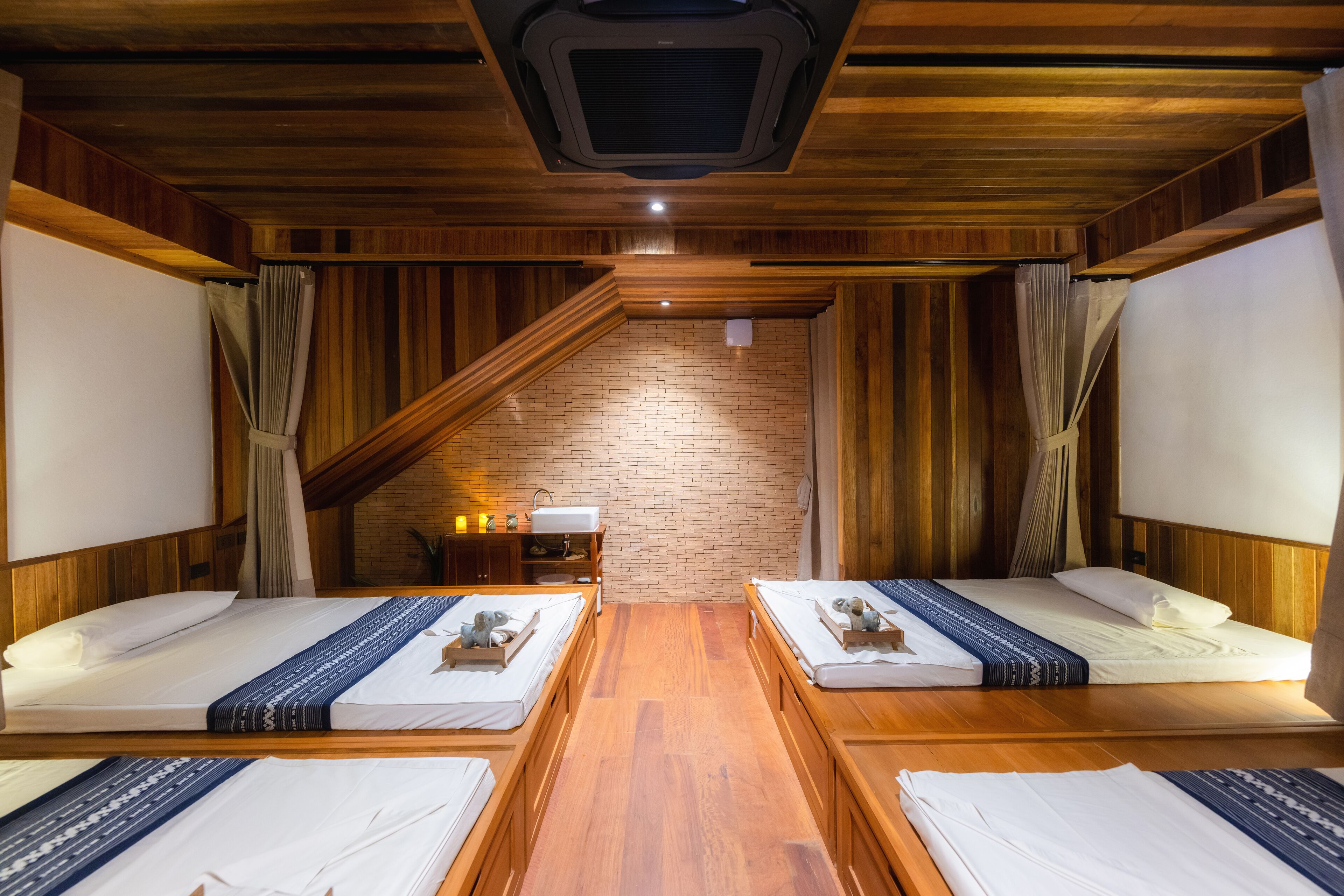 Una habitación con dos camas, piso de madera, pared de ladrillo y techo inclinado con una rejilla de ventilación.