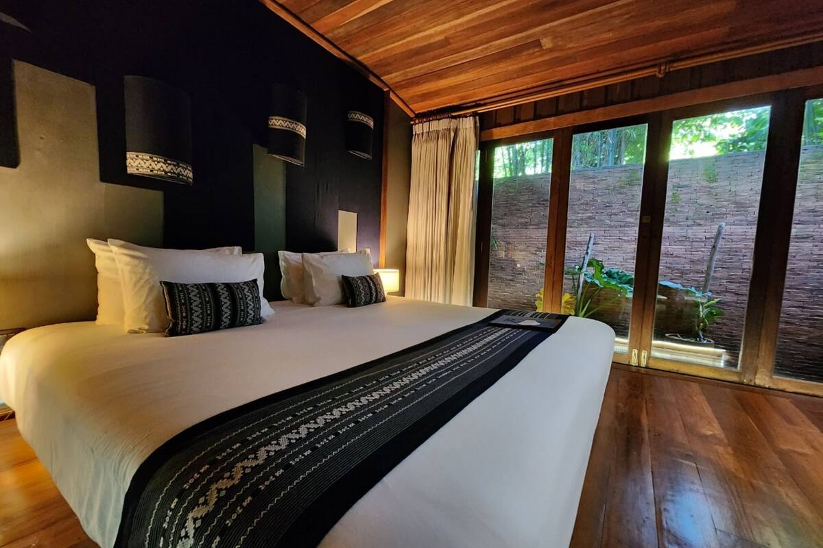 Een groot bed met witte lakens en gestreepte kussens, een houten plafond en uitzicht op groen door grote ramen.