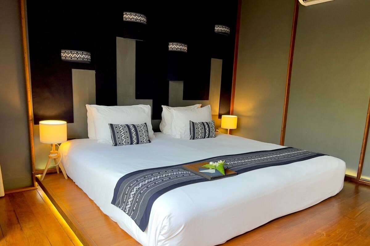 Een net opgemaakt bed met witte lakens en decoratieve kussens, een nachtkastje met een lamp, en een decoratief paneel aan de muur.