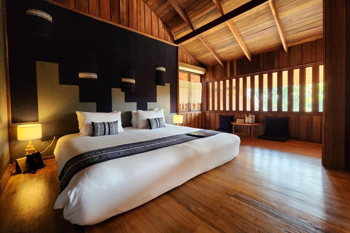 Een groot bed met witte lakens en gestreepte kussens in een houten kamer met een hoge plafond en grote ramen.