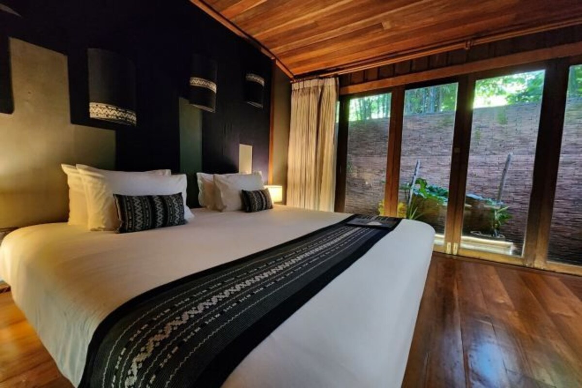 Un grand lit avec du linge de lit blanc et un couvre-lit noir à motifs, un plancher en bois et une vue sur la verdure par de grandes fenêtres.