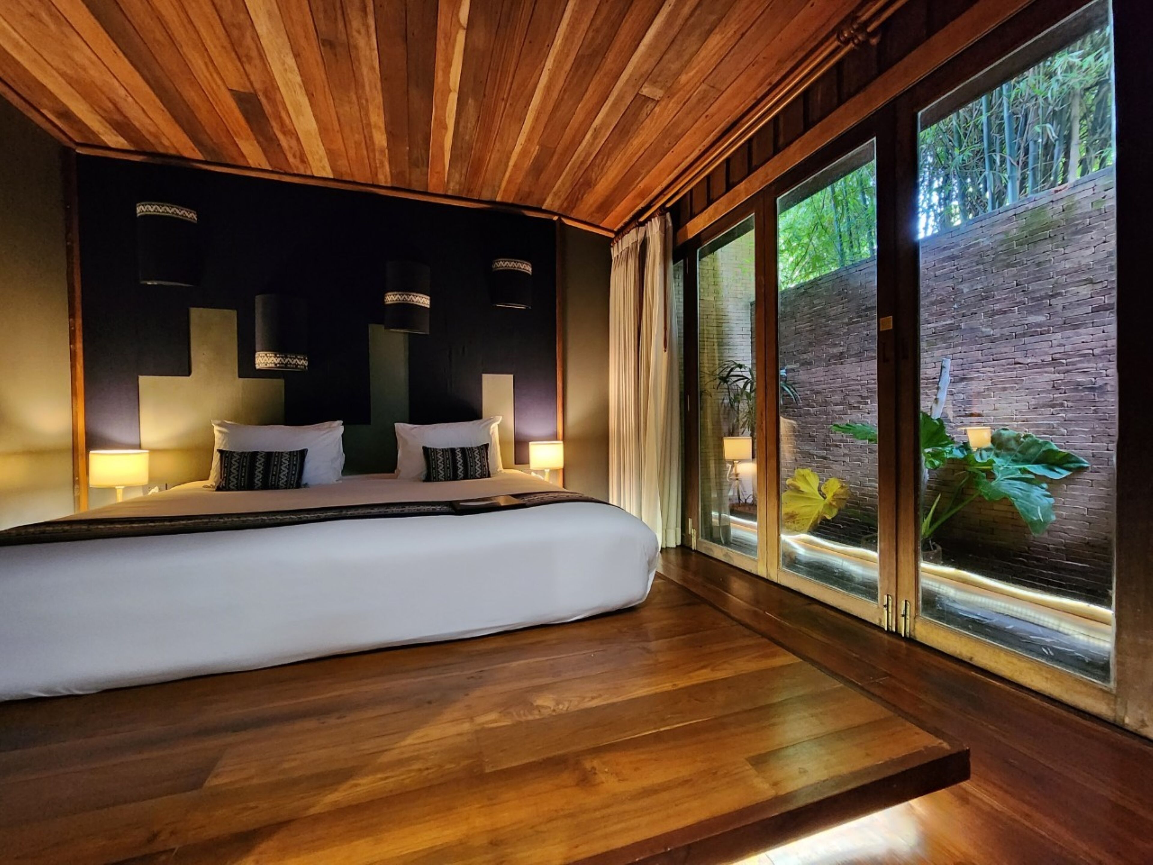 A modern bedroom with a large bed, wooden ceiling, and a view of a brick wall and greenery outside.