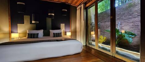 A modern bedroom with a large bed, wooden ceiling, and a view of a brick wall and greenery outside.
