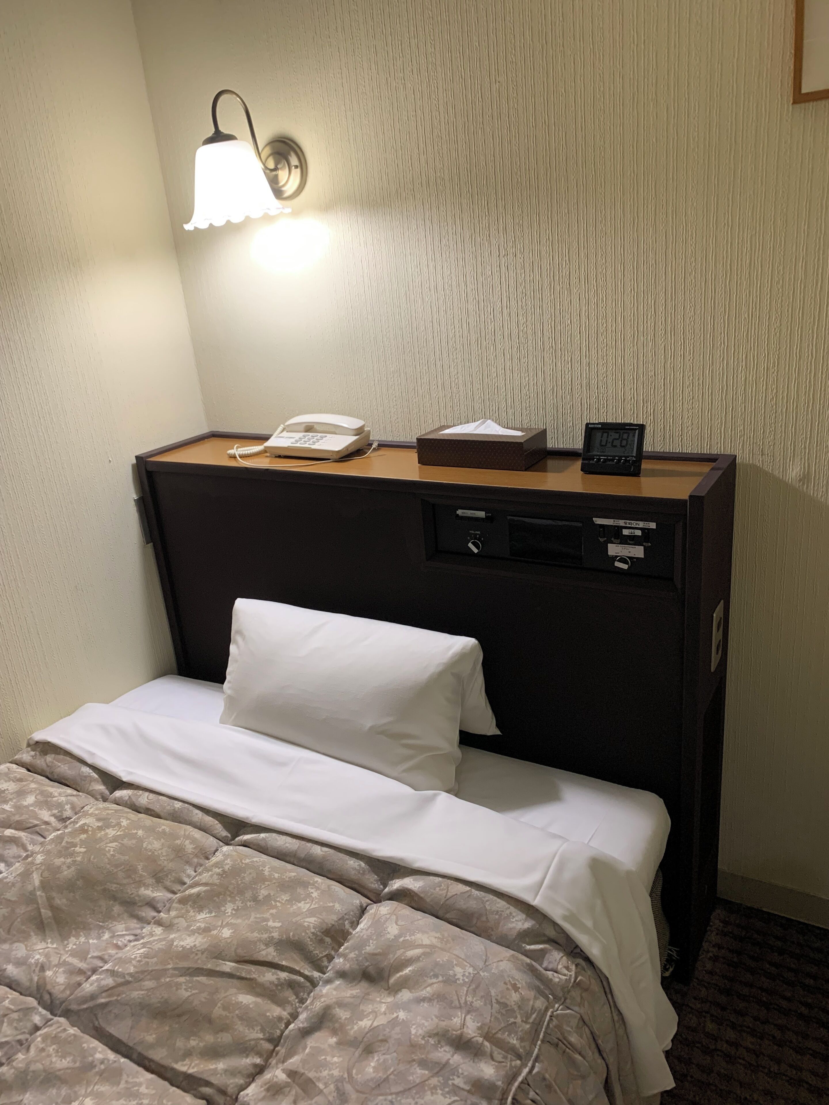 A hotel room with a bed, a bedside table with a phone, a lamp, and a digital clock.