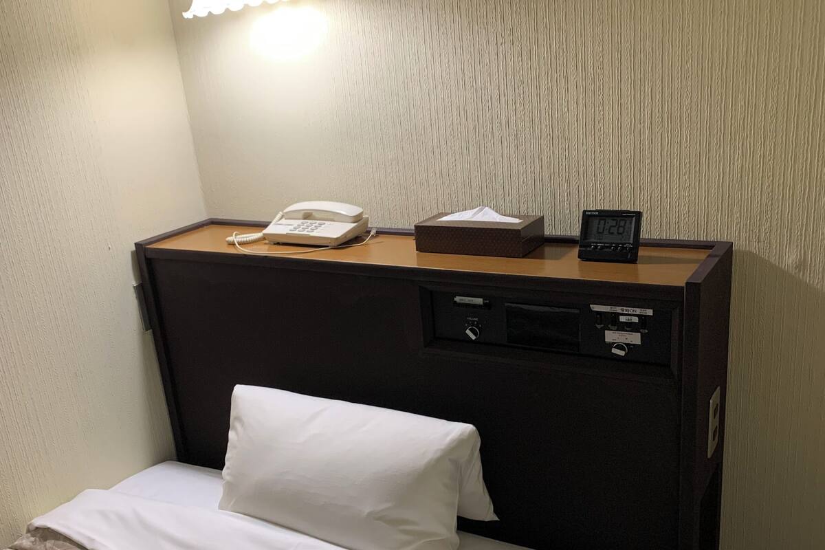 Habitación de hotel con cama, mesita de noche con teléfono, lámpara y reloj digital.