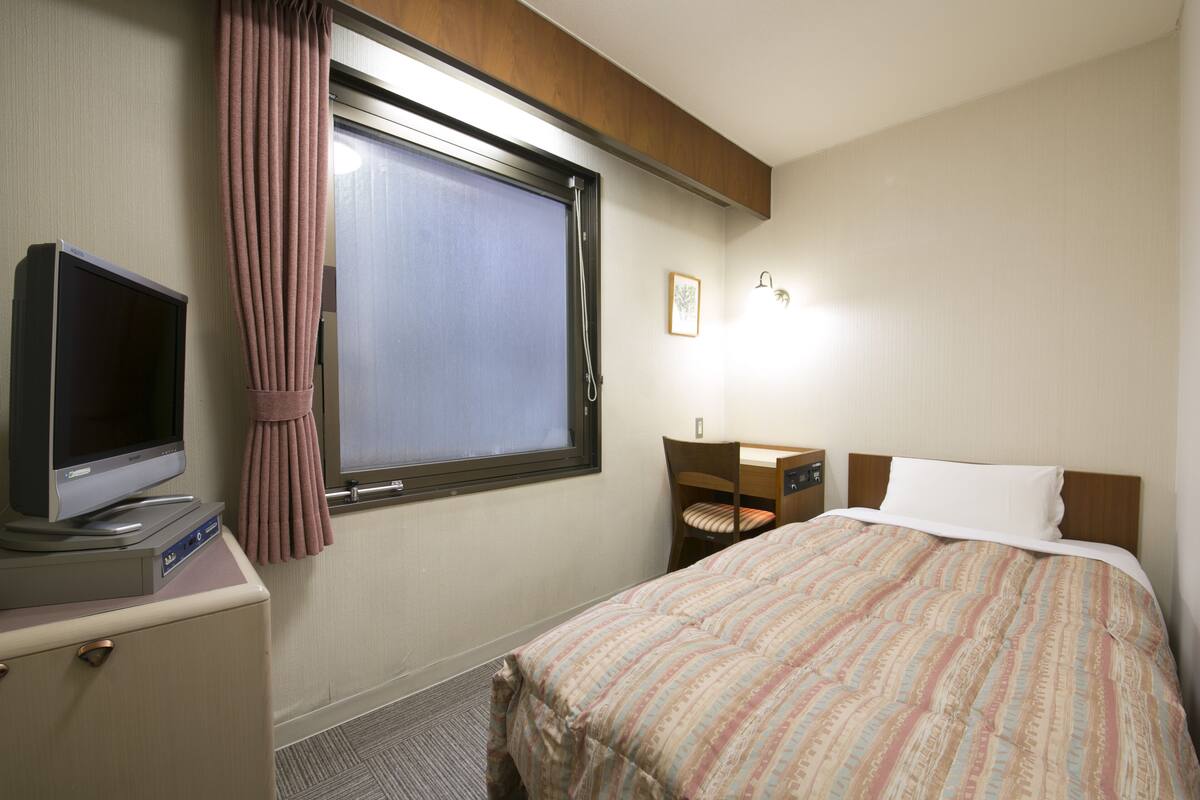Una habitación de hotel con cama, escritorio con silla, televisor en mueble y ventana con cortinas.