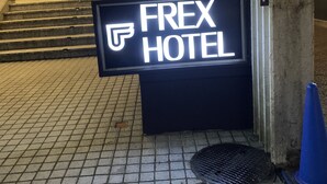 Señal del Hotel Frex, de diseño moderno, ubicada en la entrada de un edificio con pavimento de baldosas y un bolardo metálico cercano.