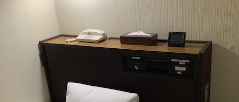 Quarto de hotel com uma cama, uma mesa de cabeceira com telefone, caixa de lenços e relógio digital.