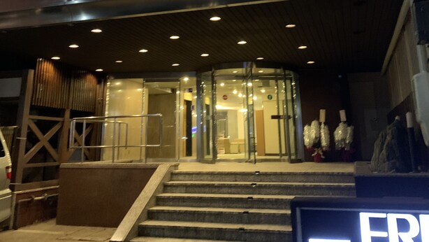 Entrada de un hotel de varios pisos con fachada de vidrio, una escalera que conduce a la entrada y un cartel con un logotipo.