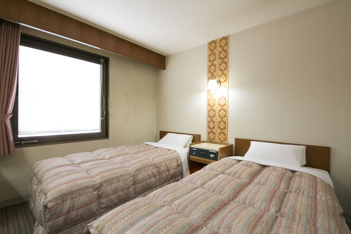 Habitación de hotel con dos camas, una ventana, una mesita de noche y una lámpara fijada a la pared.