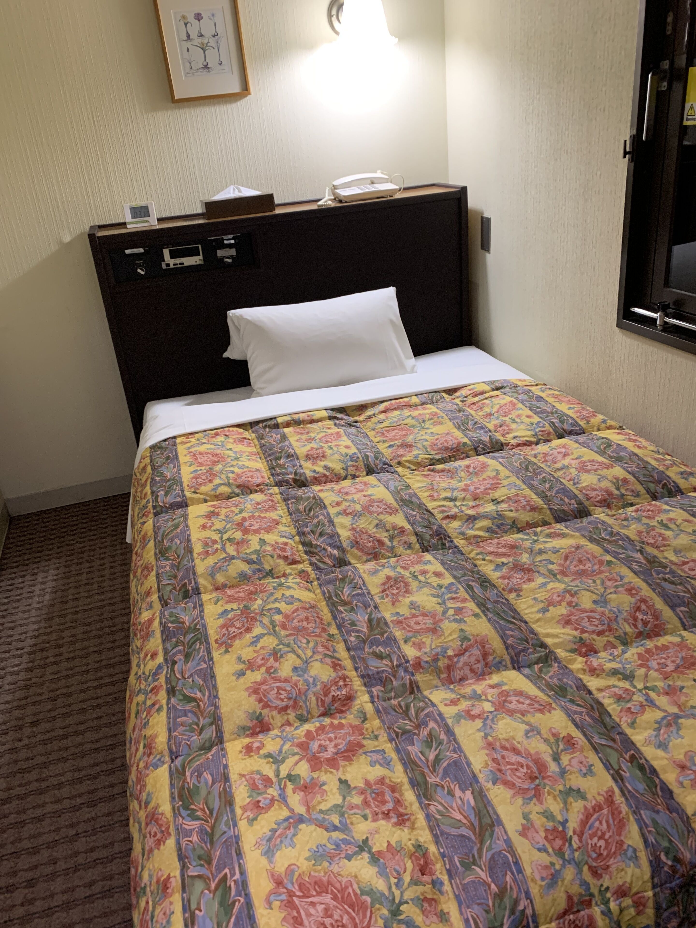 A hotel room with a bed, a bedside table with a lamp, a clock, and a framed picture on the wall.