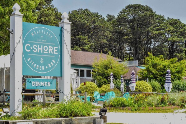 Un área de comedor exterior bien amoblada con un letrero de C*SHORE Kitchen + Bar, rodeada de follaje y asientos al aire libre.