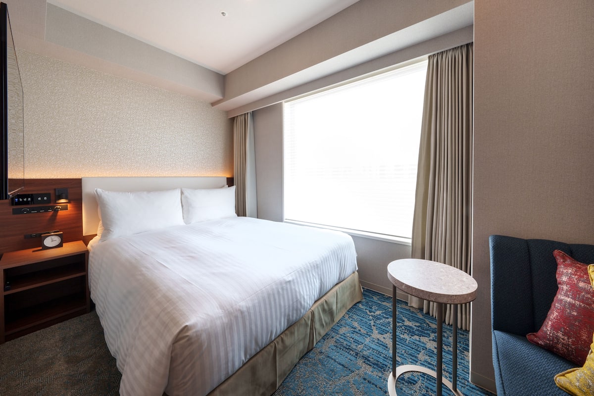 A hotel room with a large bed, a small table, a sofa, and a window with curtains.