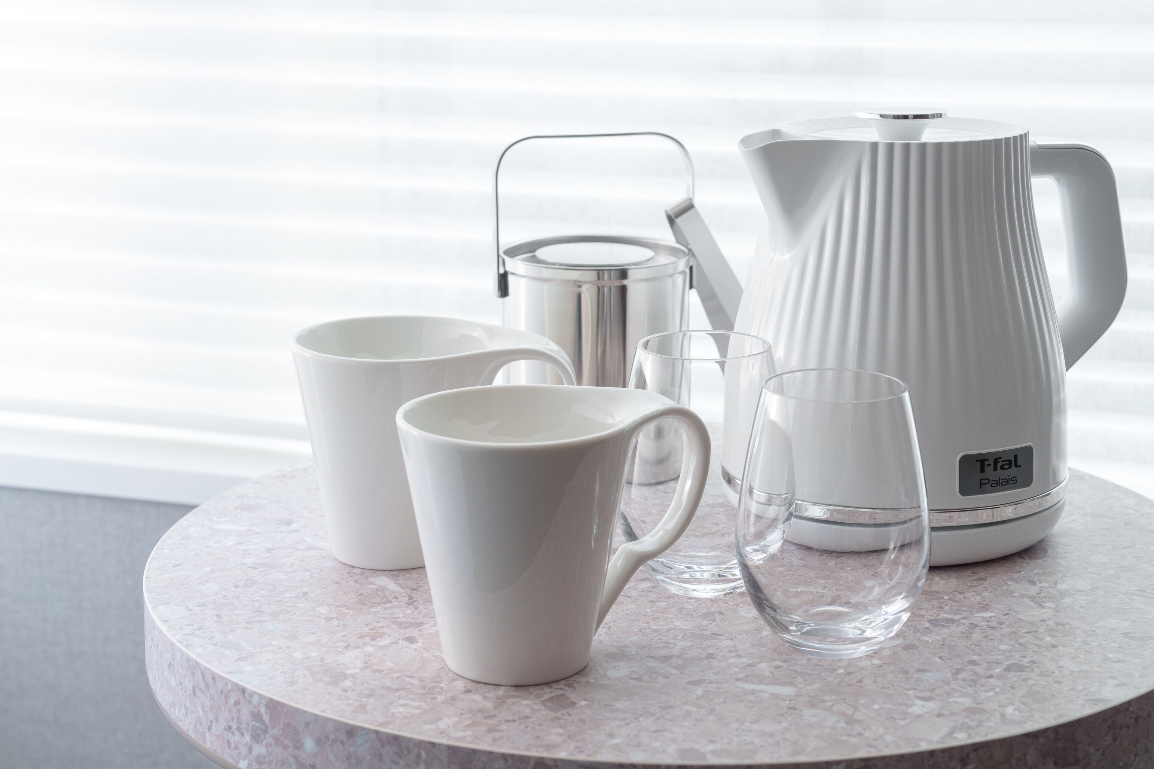 A set of white ceramic mugs, a glass, and a stainless steel coffee pot on a marble table.