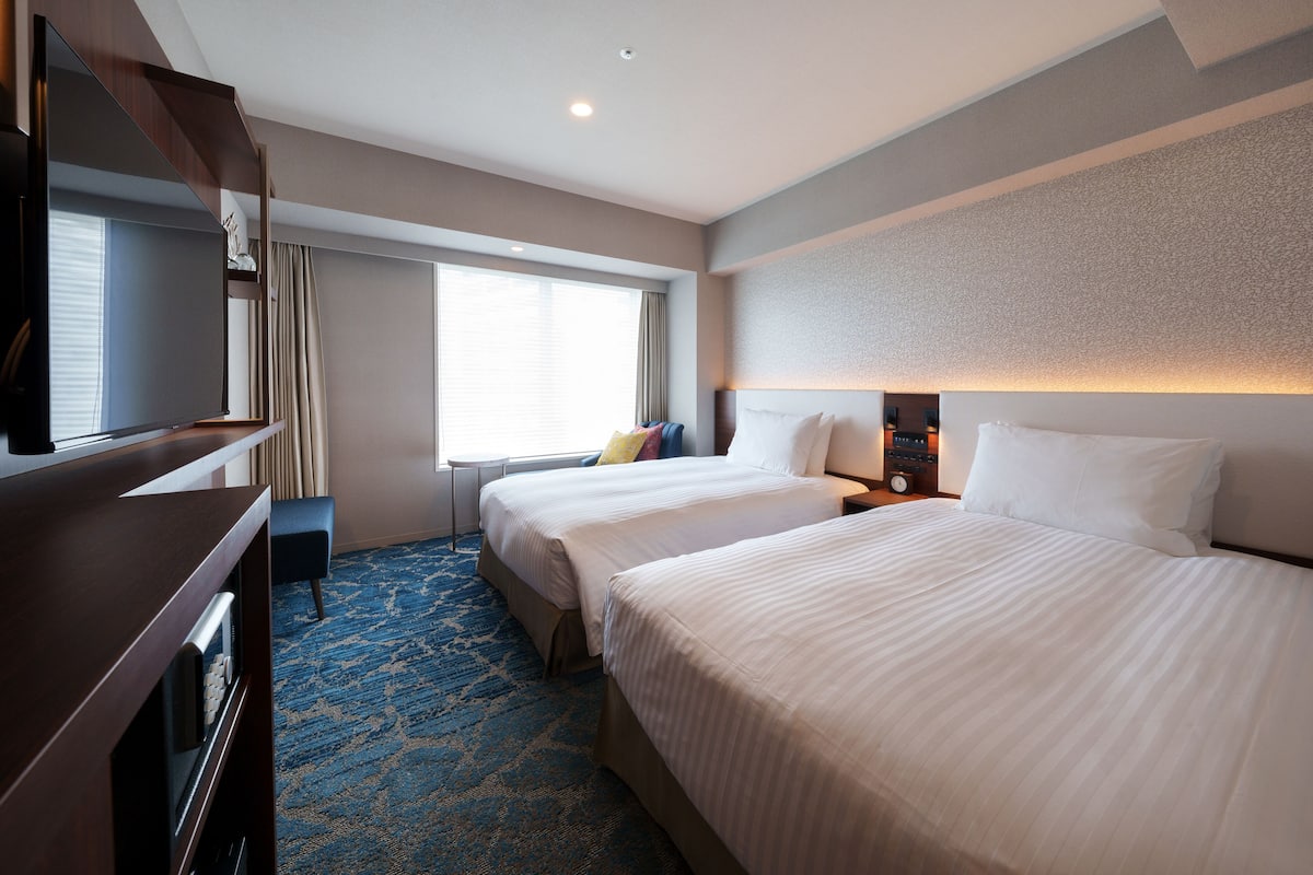 A hotel room with two beds, a TV, a desk, and a large window.